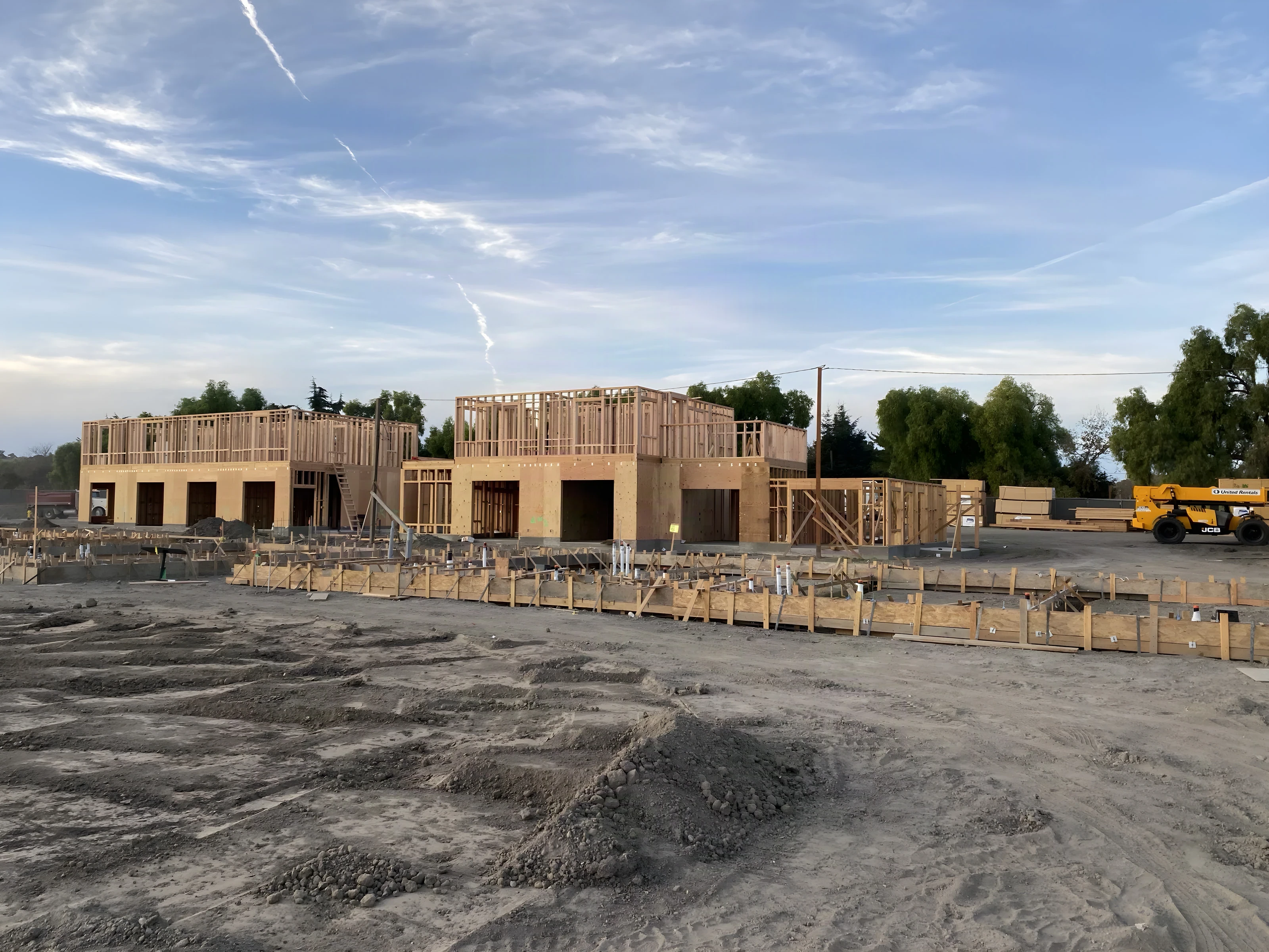Ground-up wood framing rising across a graded construction site beside mature trees