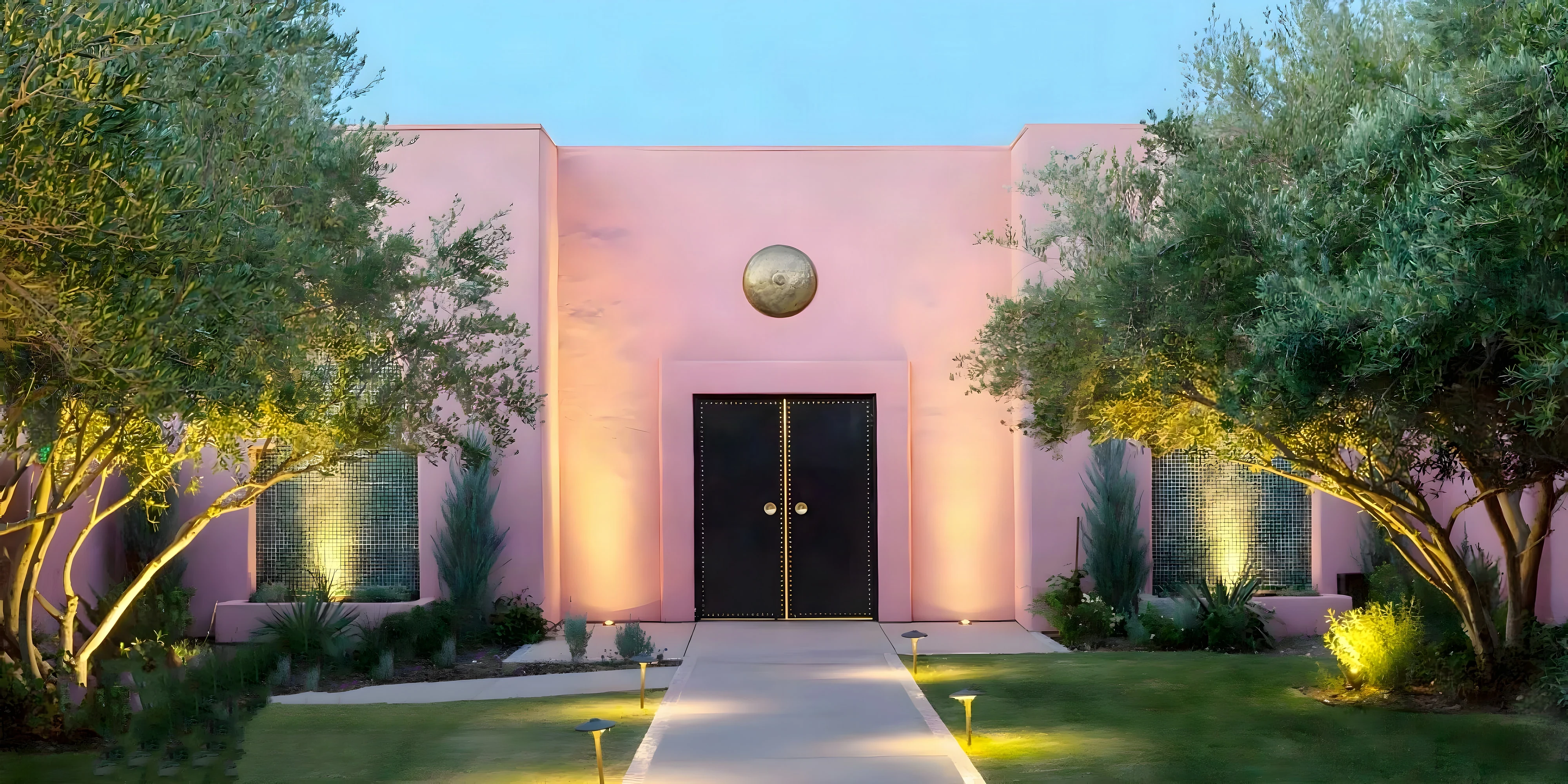 Pink stucco spa entrance with black studded doors between uplit olive trees at dusk in Indian Wells