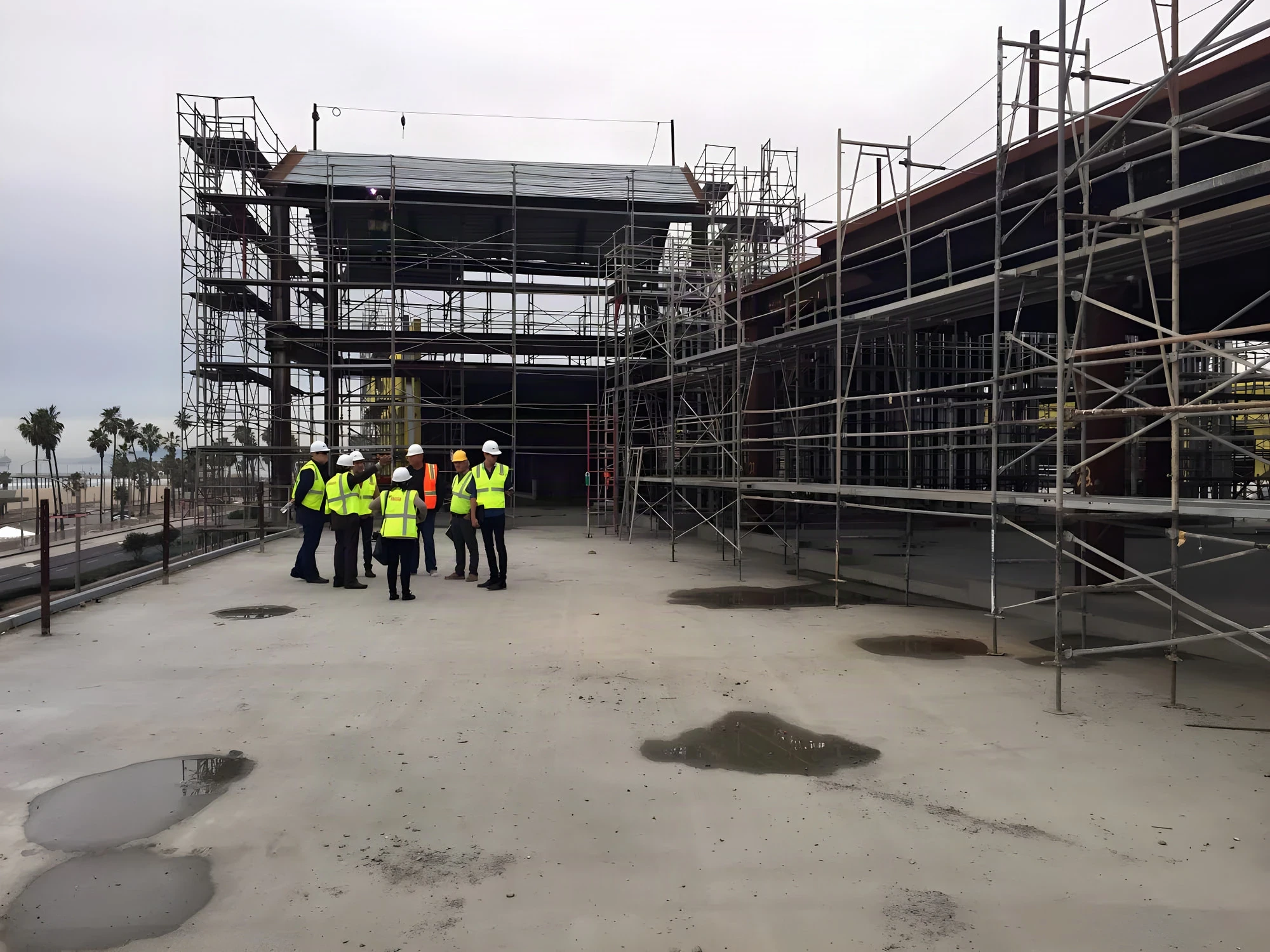Hi-vis crew in hard hats gathered on a concrete deck beside scaffolding, with the pier and palms beyond