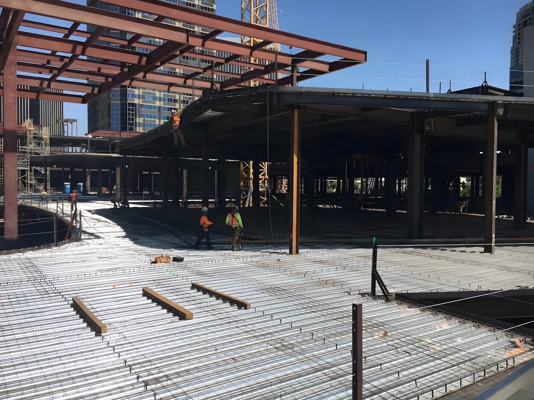 Crew in hi-vis vests crossing rebar-laid metal decking beneath a downtown steel frame